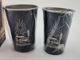 Two black cups with white designs on a white surface, with a cardboard box in the background.